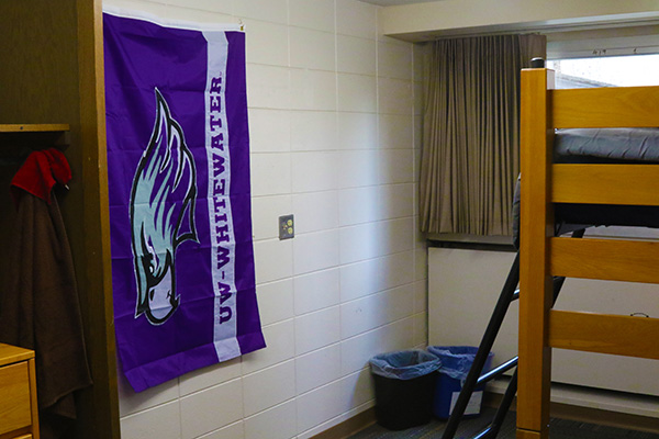 A dorm room. The left side has 2 trash cans and a UW Whitewater flag, and the right side has a lofted bed.