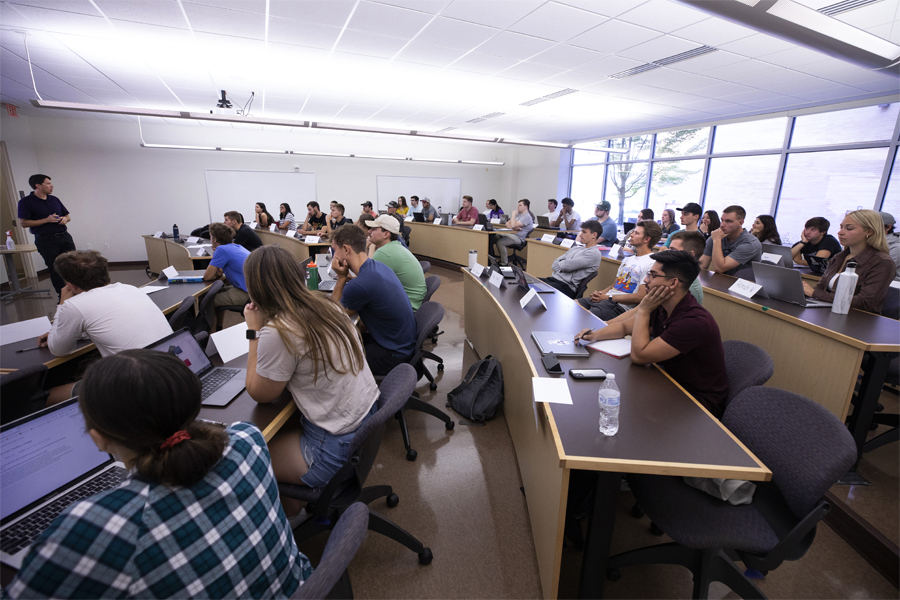 Students sit at desks in a large classroom.