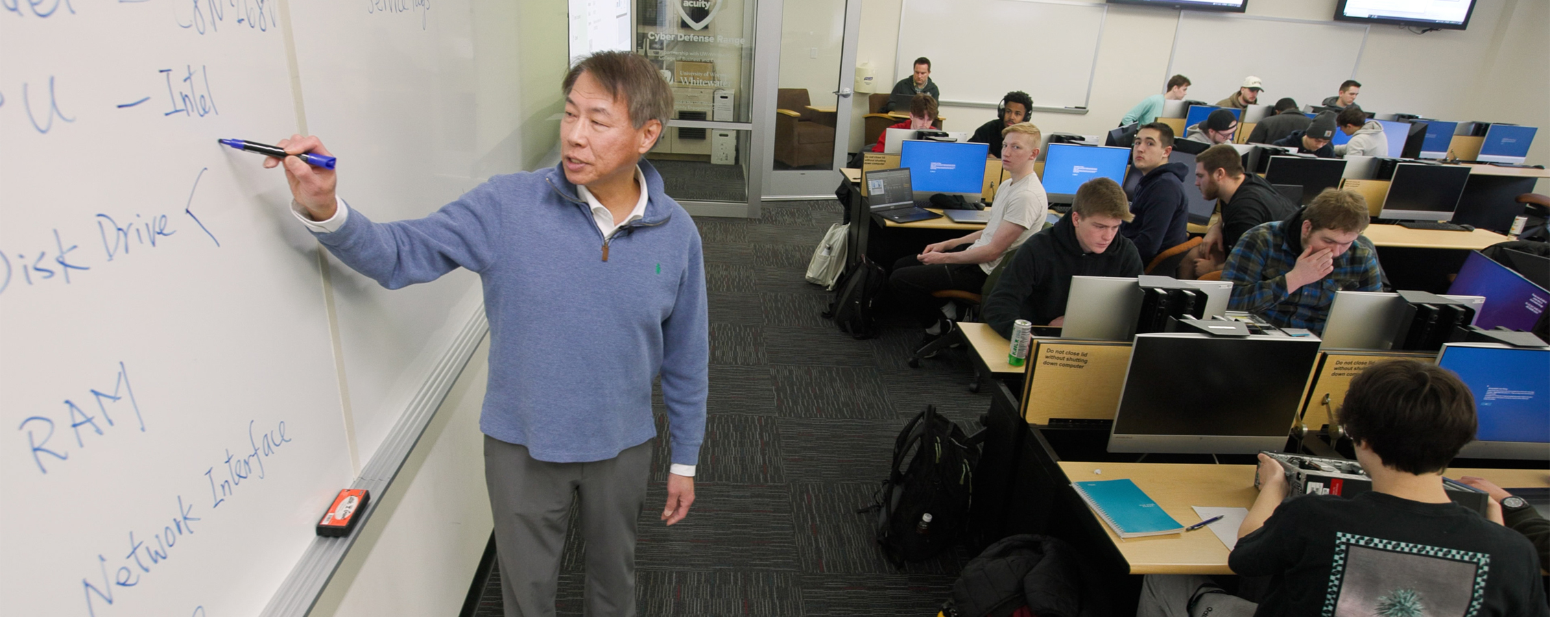 Roger Yin wears a blue sweater and teaches in front of a class as he writes on a whiteboard.