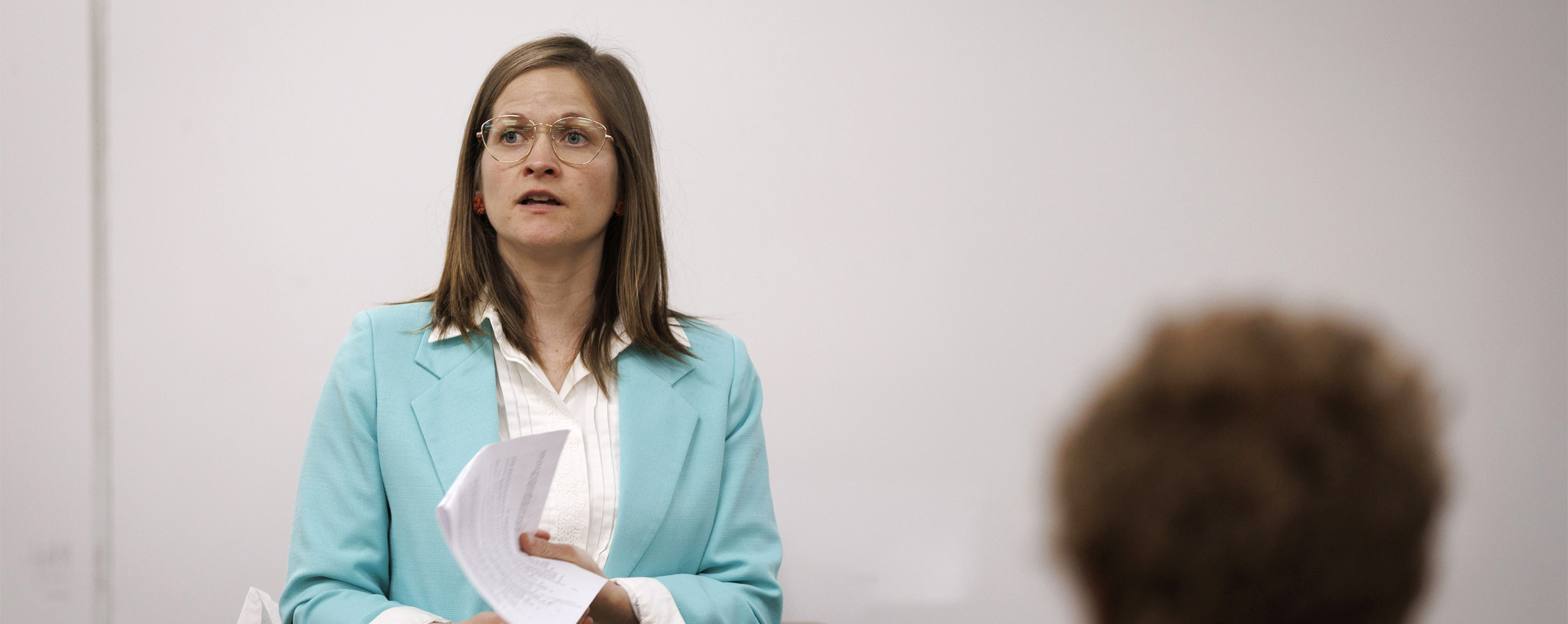 Kelly Jensen, wearing a teal blazer, speaks at the front of class while holding a piece of paper.
