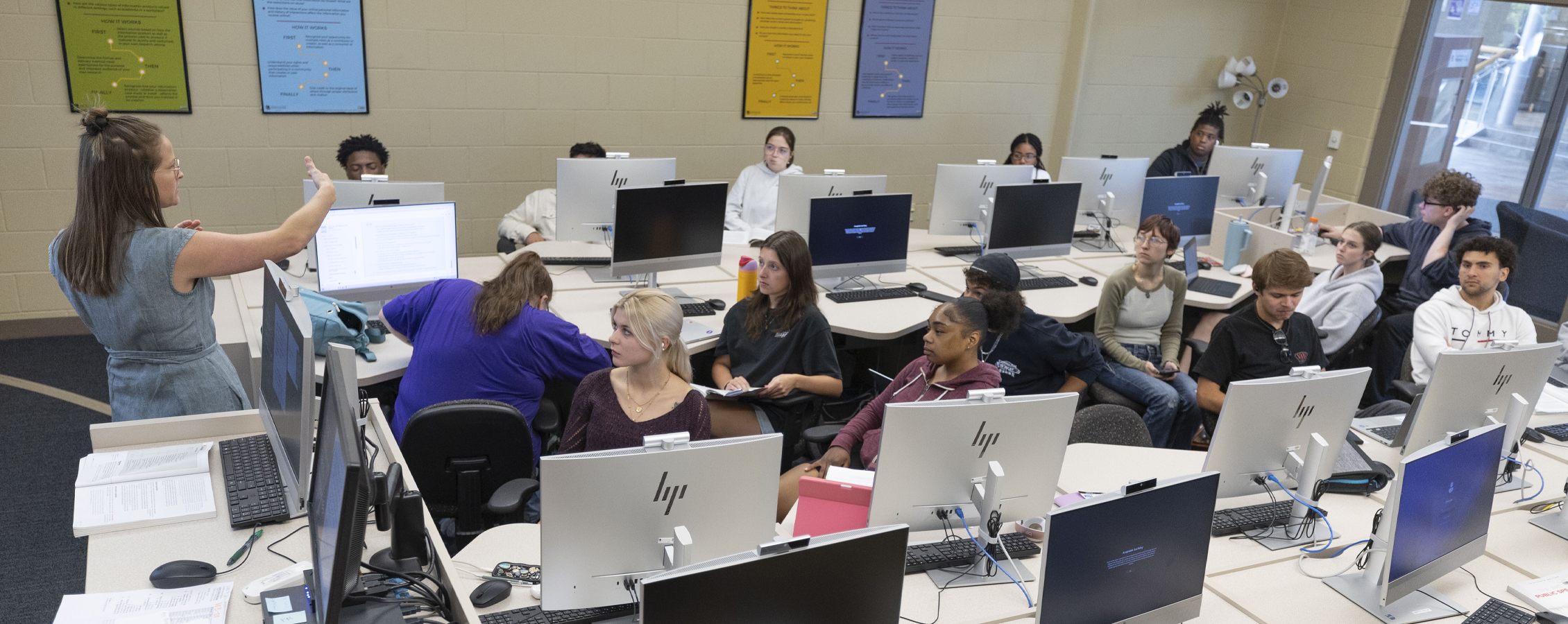Kelly Jensen uses her hands as she speaks at the front of class while students sit at tables with desktop computers.
