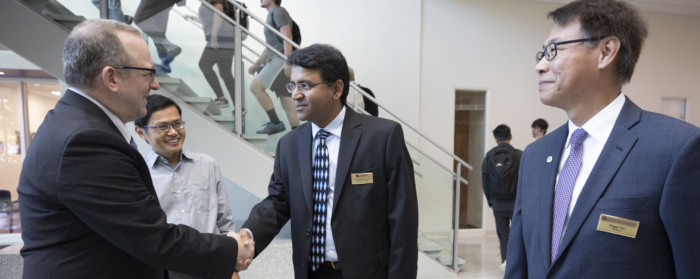 Roger Yin, dressed in suit and tie, stands to the side and smiles as two people shake hands.