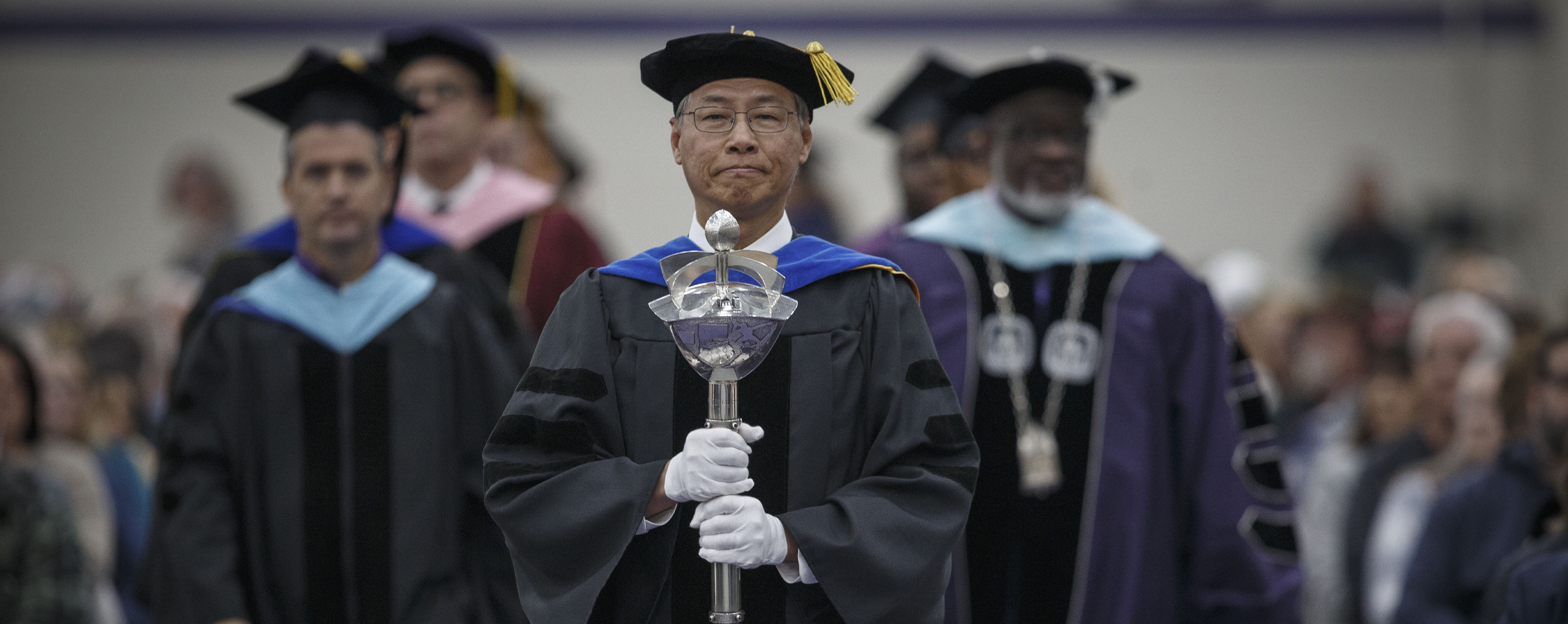 Roger Yin, dressed in academic regalia, walks down the aisle during commencement holding the ceremonial mace.