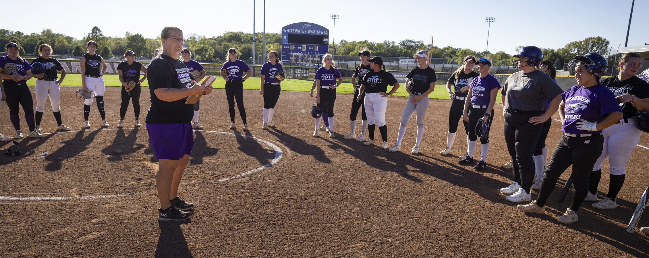 Brenda Volk stands in the infield of a ball diamond, surrounded by her players in softball uniforms.