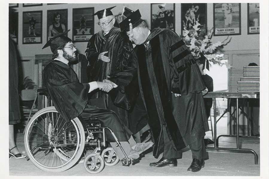 A black and white photo of a person in a wheelchair crossing the stage at graduation in cap and gown.