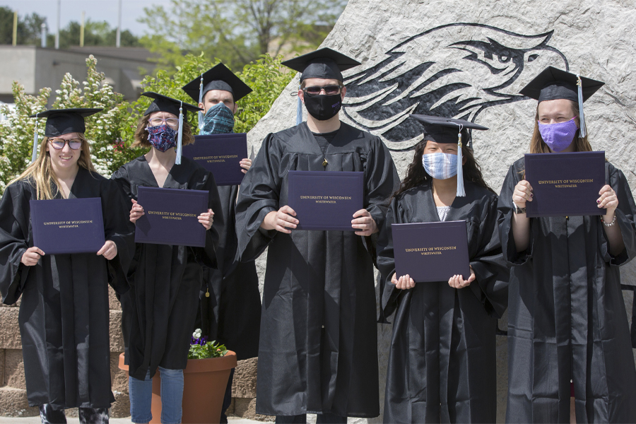 Students stand with diplomas in front of a boulder with a Warhawk head engraved on it.