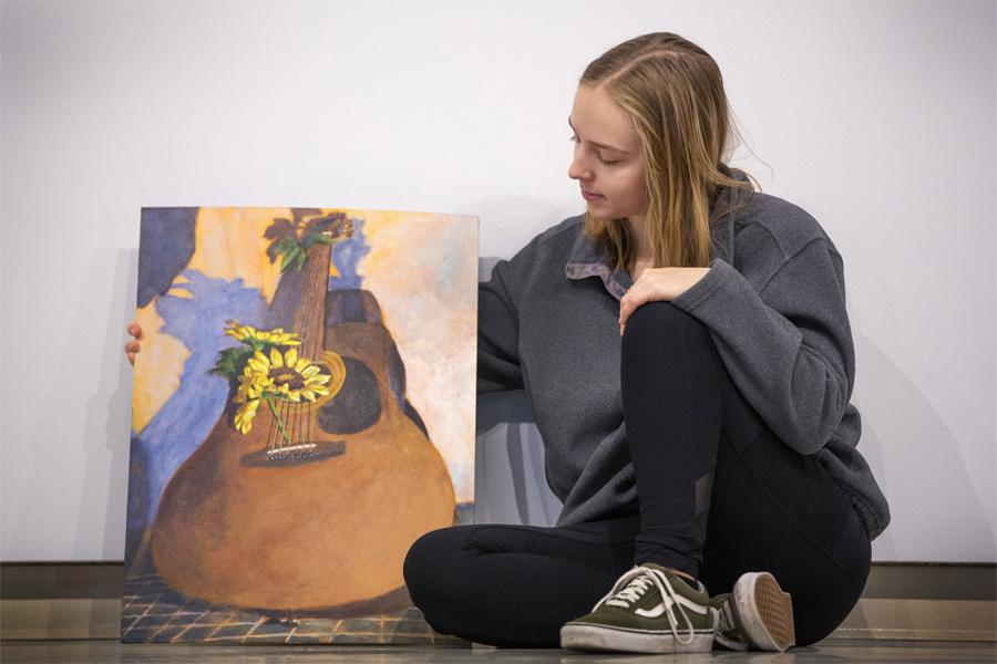 Emma Siskoff sits next to a painting on a guitar.