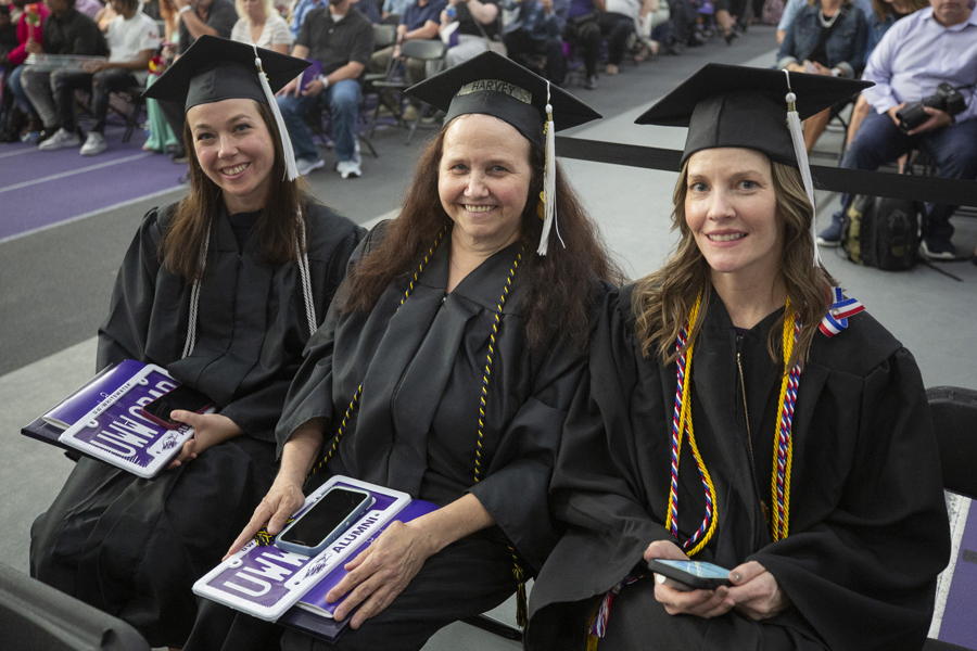 Three adults wearing caps and gowns sit together at graduation.