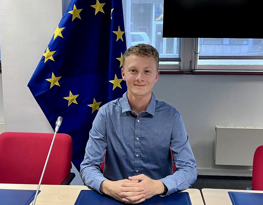 Dylan Riley sits at a desk with the European Union flag behind him, which is a blue flag with yellow stars.