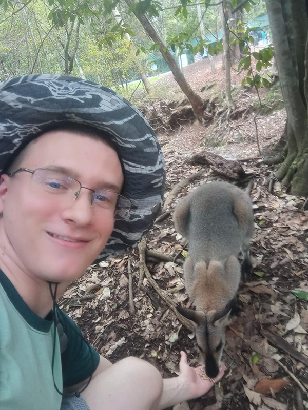 A student wears a camoflauge hat and feeds a kangaroo out of his hand.