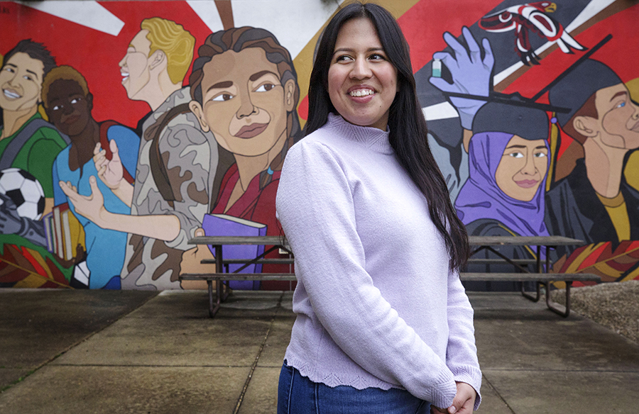 Maria Pacheco stands in front of a colorful mural with her hands folded and looking off to the left.