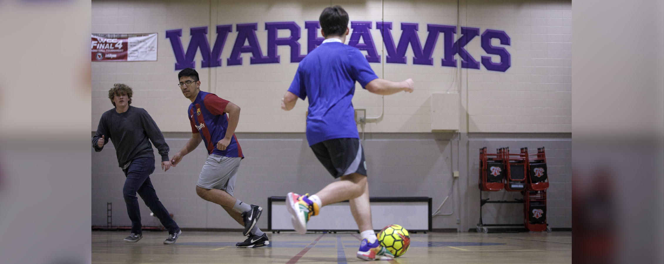 A student dribbles a soccer ball in a gym with the words Warhawks on the wall.