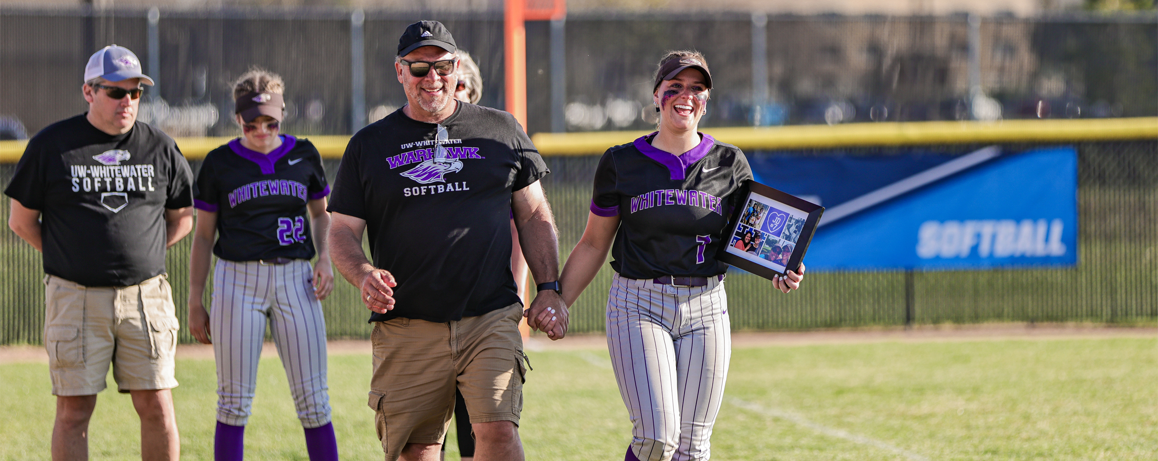 A Warhawk softball player walks hand in hand with her dad on the softball field while smiling and holding a plaque.