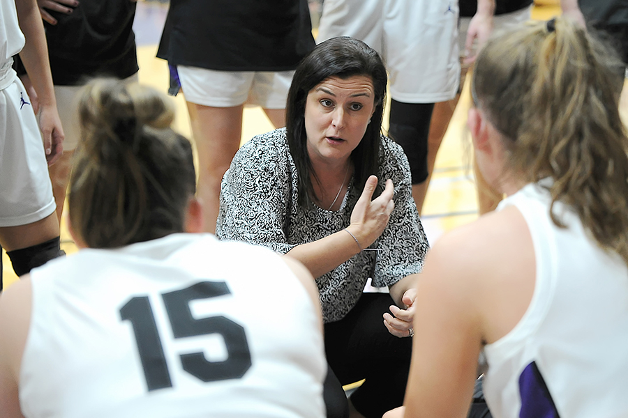 Coach Carollo kneels down to talk to her players sitting on a bench.