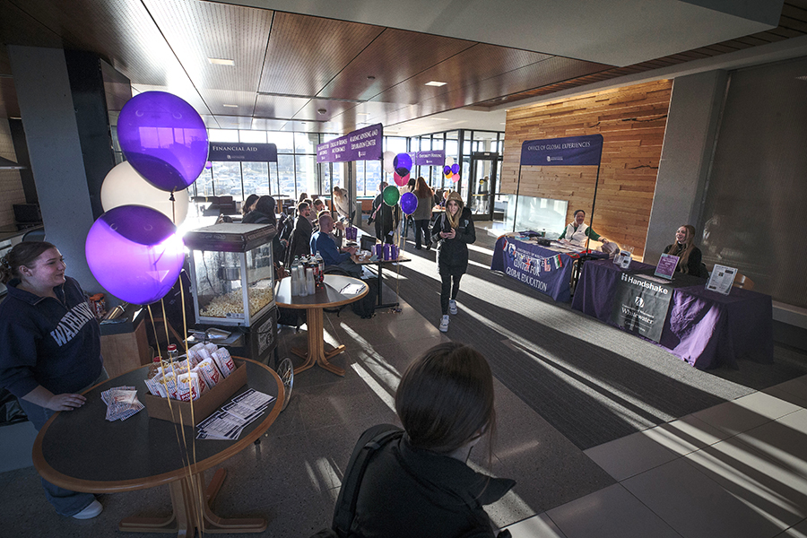 People mingle in a room with large windows and purple and white balloons.