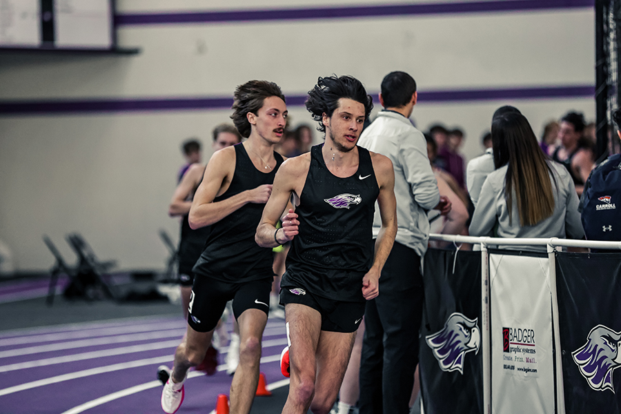 Cross country runners in black jerseys run on an indoor track. 