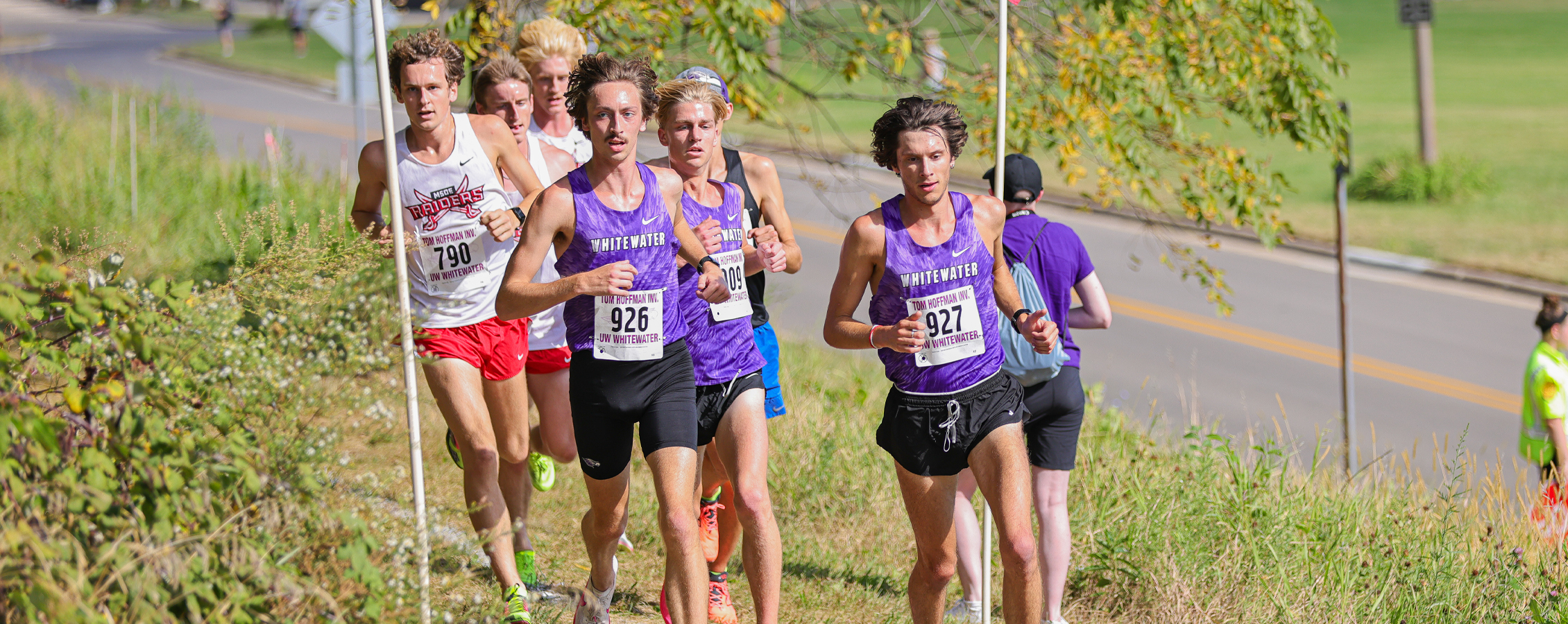 Three Warhawks in purple shirts run in the front of a pack of cross country runners who run by flags on an outdoor trail.