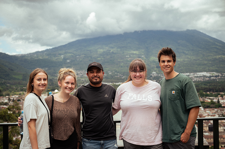 A group of five people stand together in front of an urban area with a mountain and cloud cover in the background. 