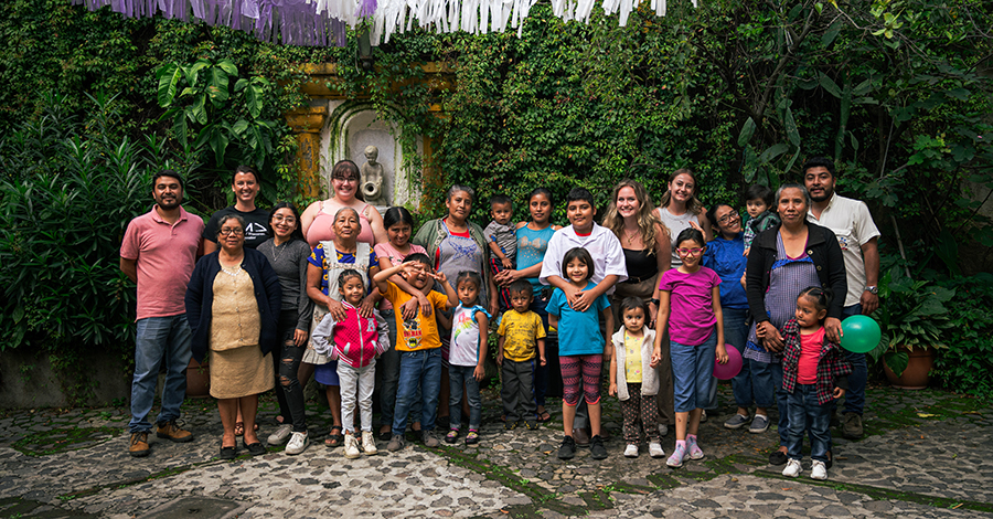 Un nutrido grupo de adultos y niños se encuentran juntos frente a una pared parcialmente cubierta de exuberante vegetación. 