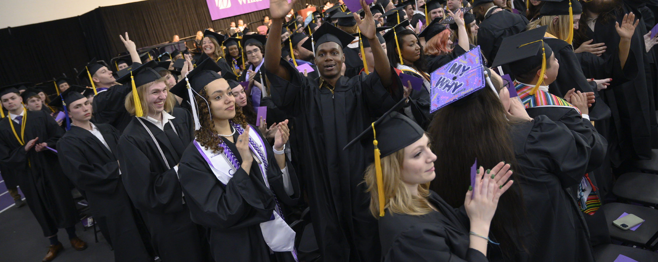 A group of graduates clap at commencement dressed in caps and gowns.