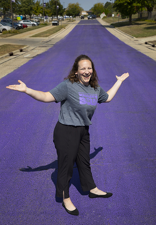 Jenny Krzyszkowski stands on the purple road with a smile on her face and her arms stretched out.