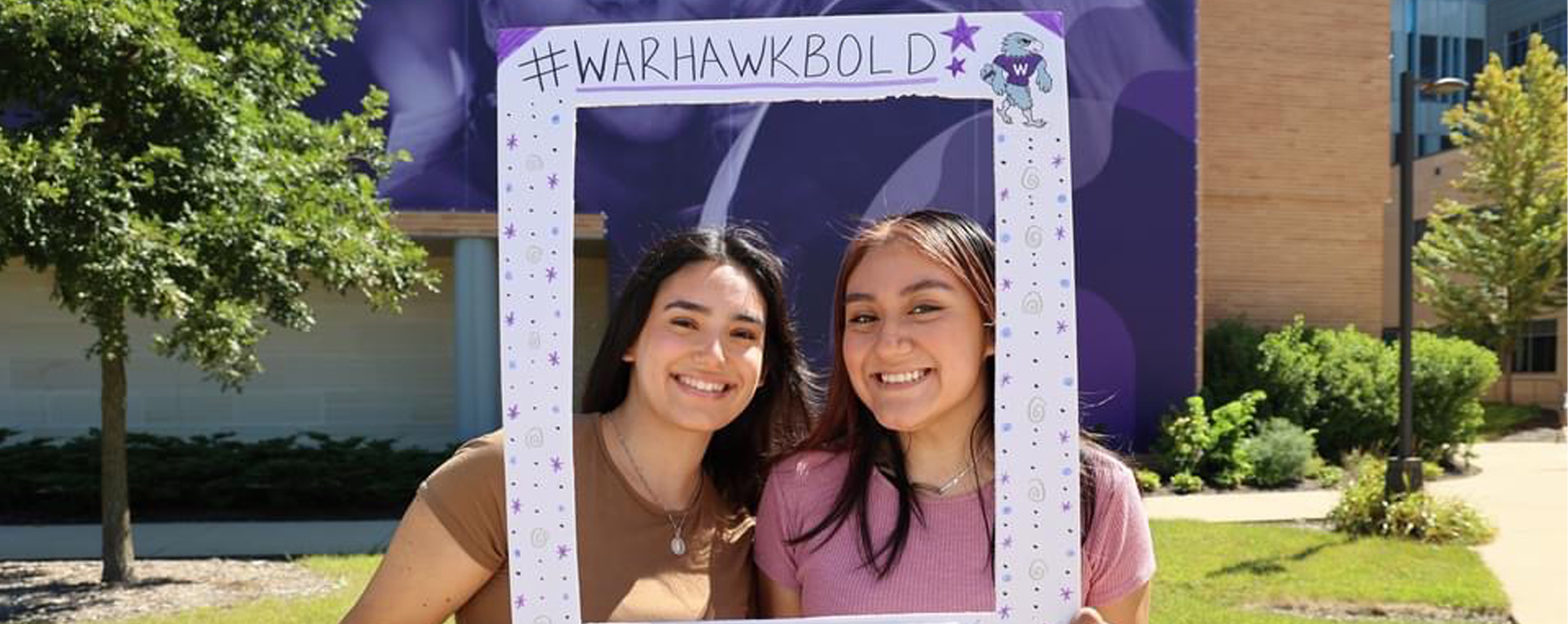 Two students pose for a photo outdoors and hold a large frame that says Warhawk Bold.