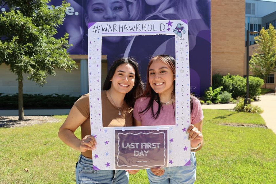 Two students pose for a photo outdoors and hold a large frame that says Warhawk Bold.