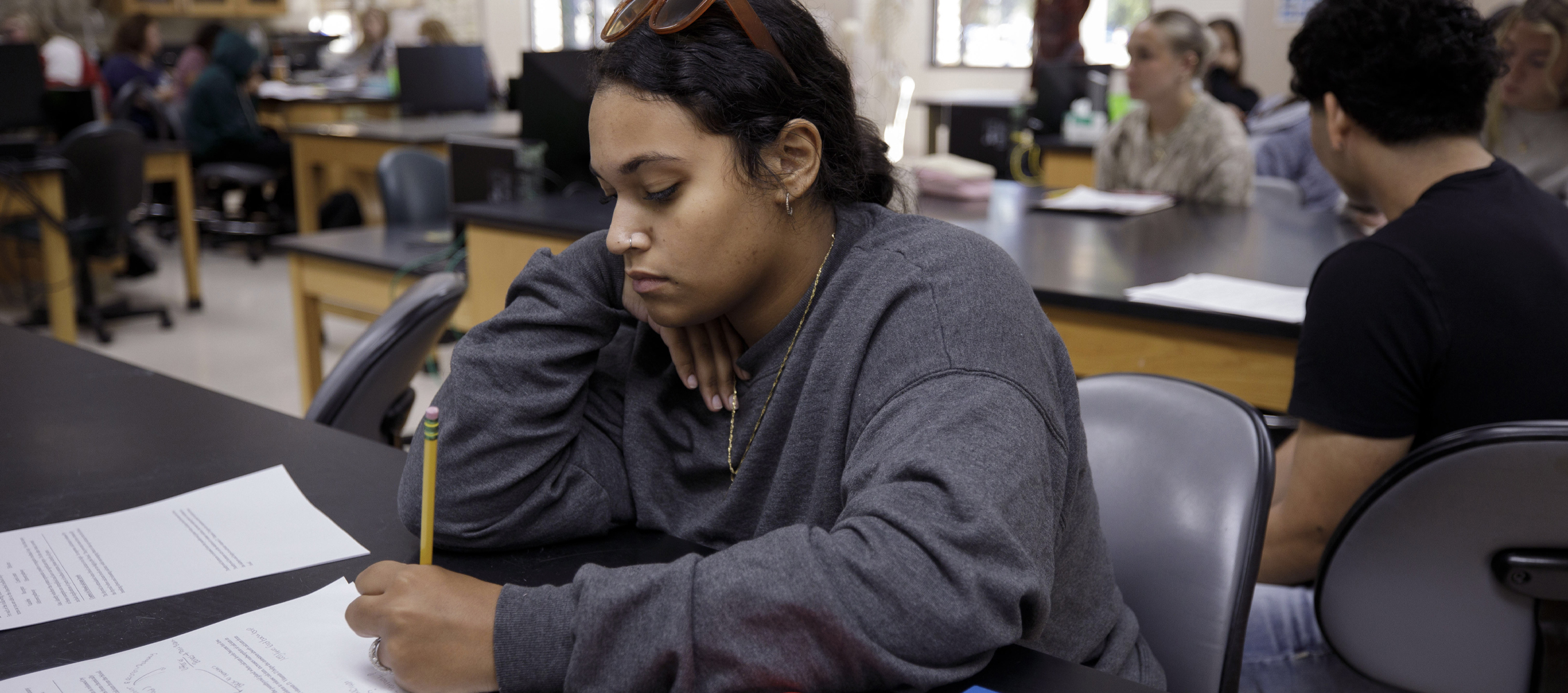 A student sits at a table and takes notes with a pencil.