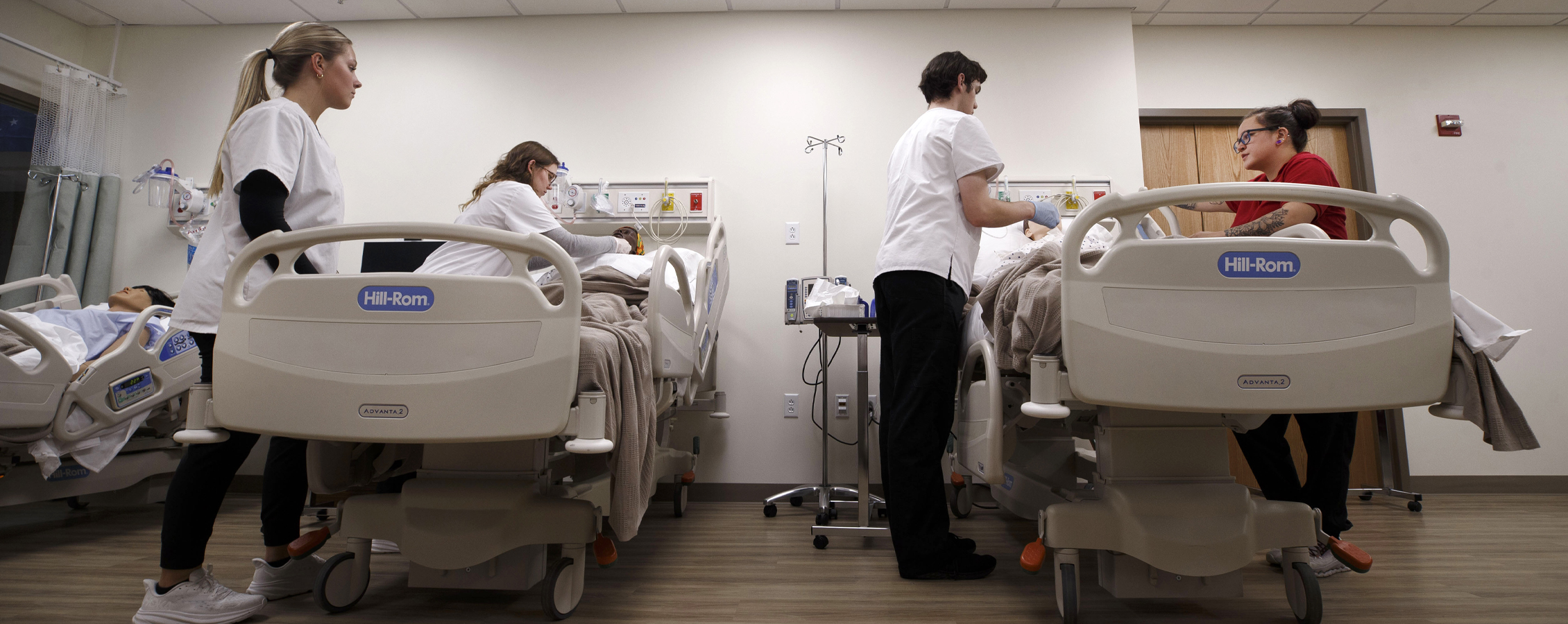 Students stand next to hospital beds with medical training manikins in the beds.