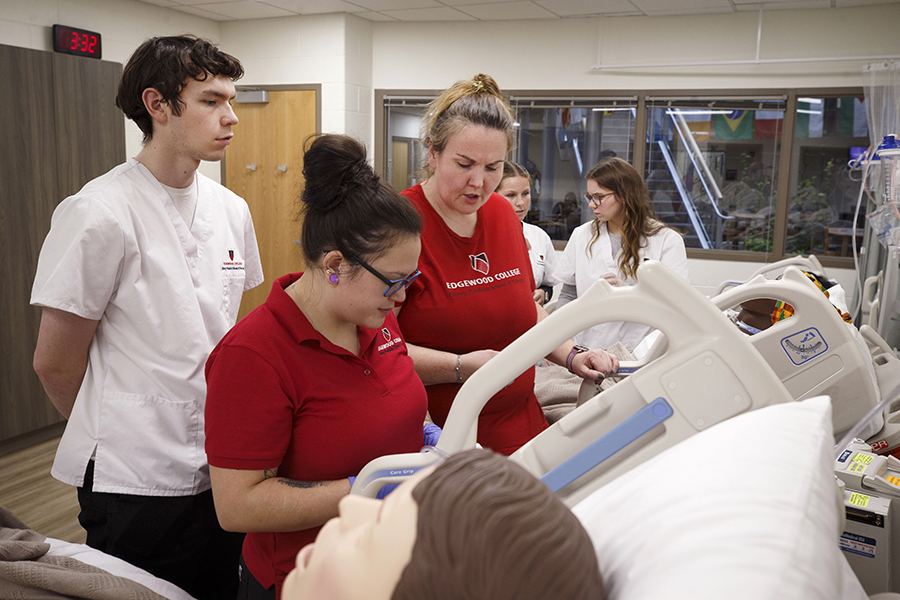 A small group of people work together as they stand next to a hospital bed with a medical training manikin in the bed.