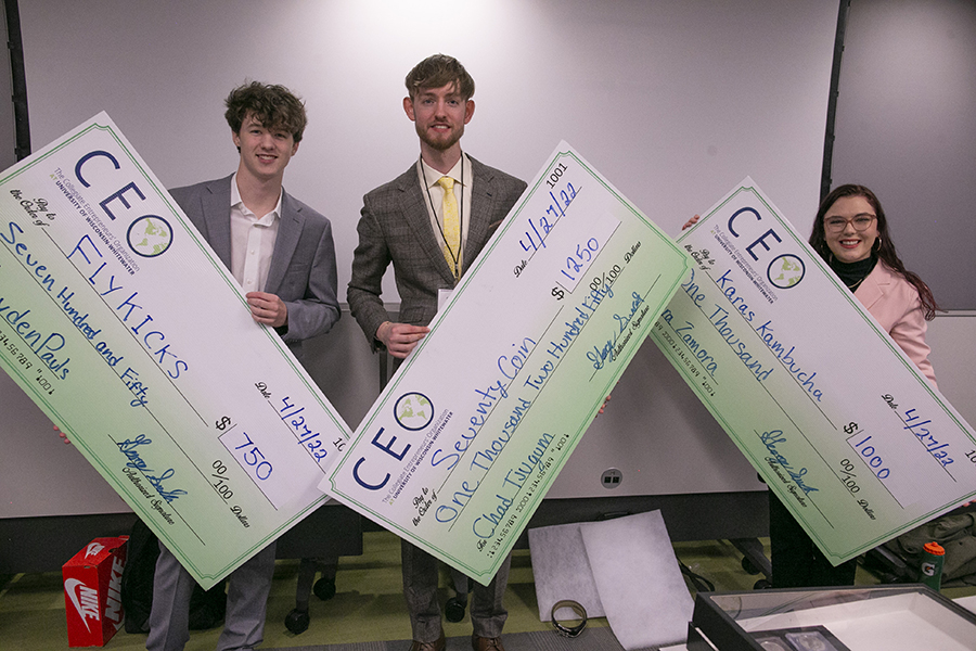 Three students stand together and pose for a photo while holding three large checks.