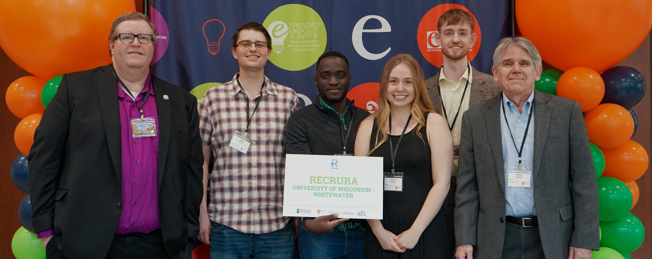 Five people pose for a photo with a plaque and orange balloons behind them.