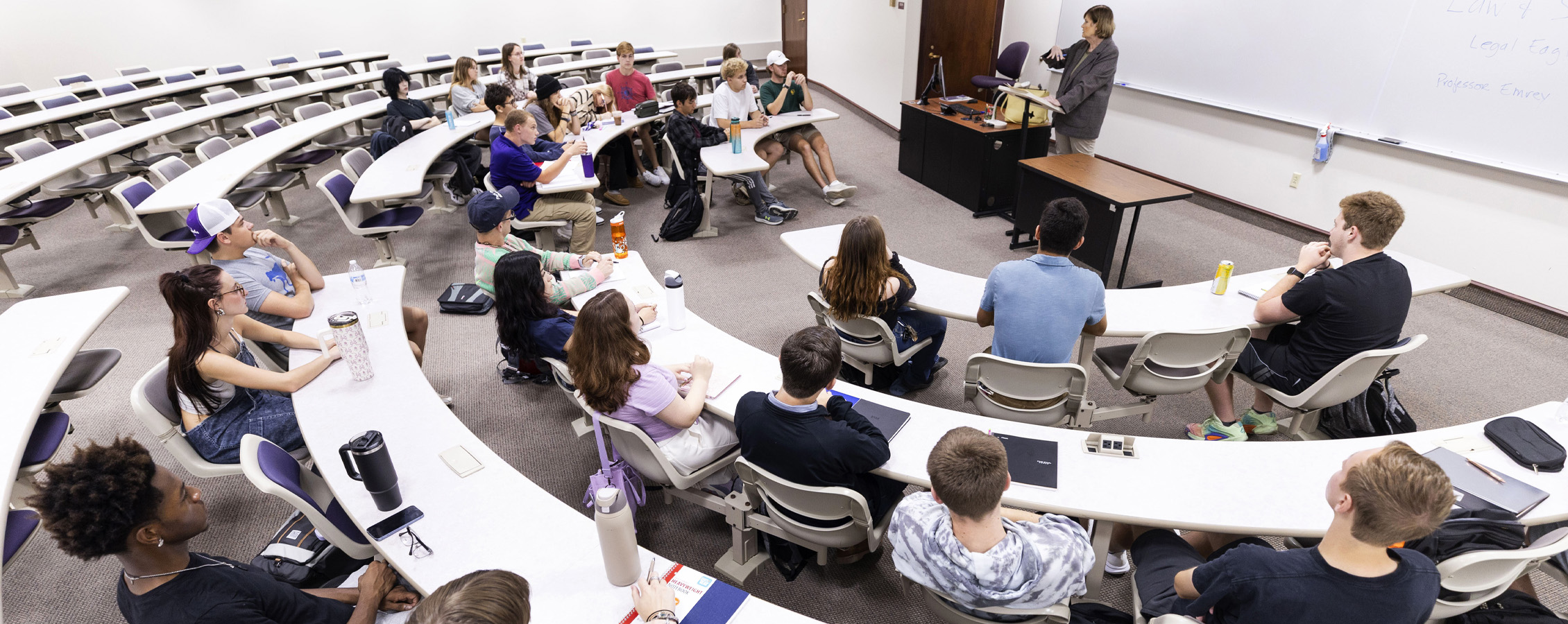 A faculty member teaches at the front of a classroom with students sitting at curved tables..