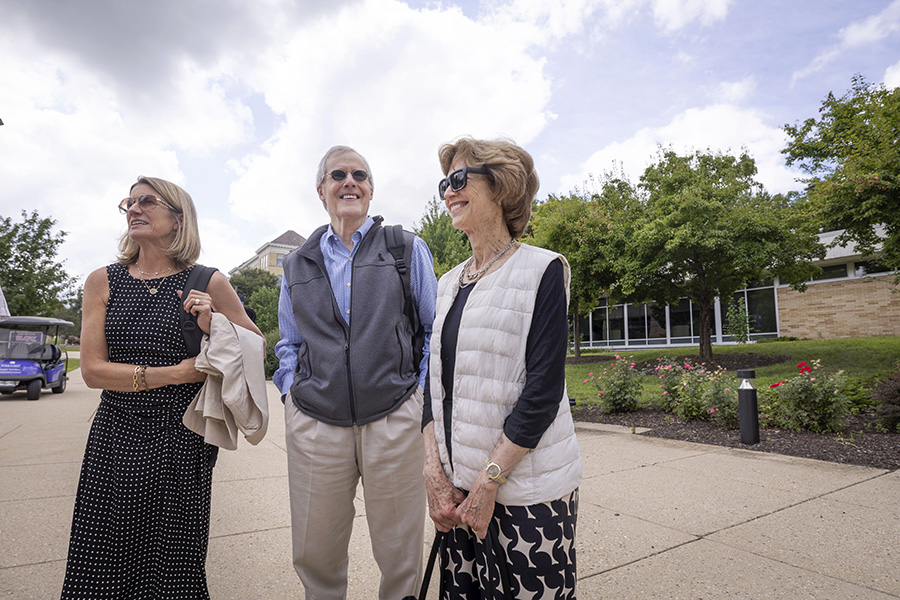 Three people stand outdoors on UW-Whitewater campus grounds with trees and flowers in the background.