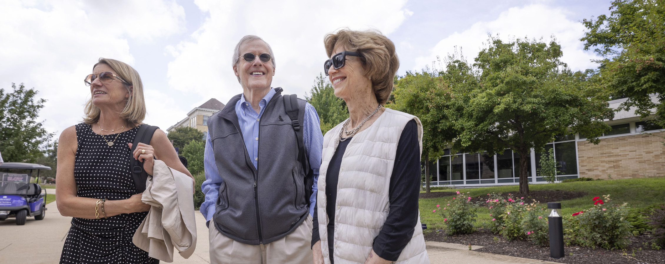 Three people stand outdoors on UW-Whitewater campus grounds with trees and flowers in the background.