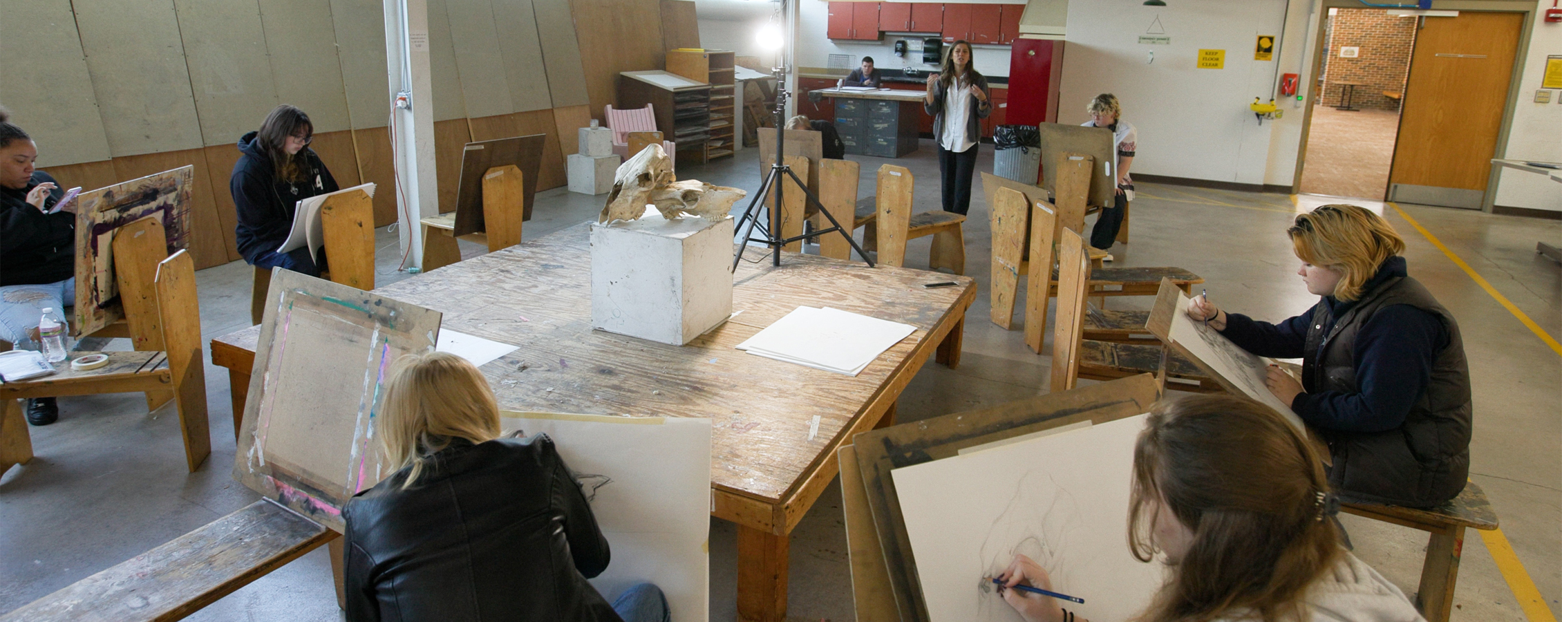 Students painting on canvases sit in a loose circle with a faculty member speaking at the front.