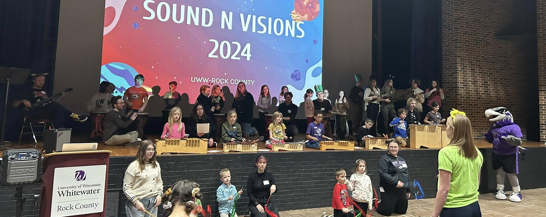 People of all ages, and Willie Warhawk, gather around a stage with a large screen that says Sound N Visions.