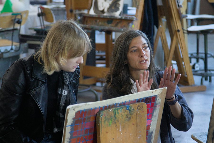 Anna Moisiadis kneels down next to a student painting on a canvas.