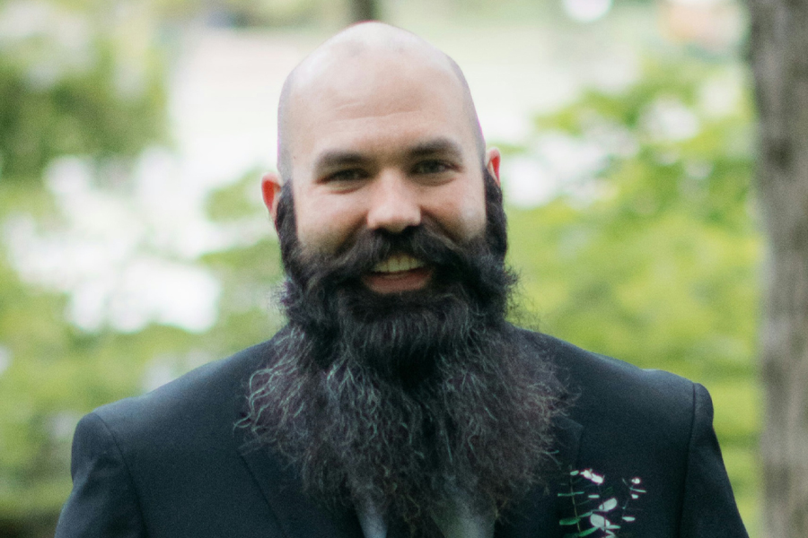 A person with a large beard stands outside and smiles at the camera.
