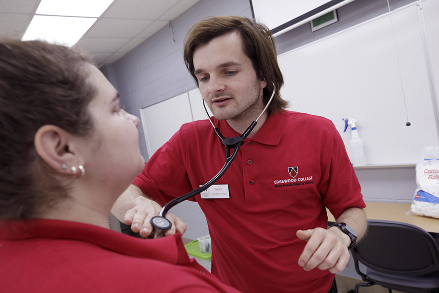 A student wearing a red shirt holds a stethoscope to a person's chest.