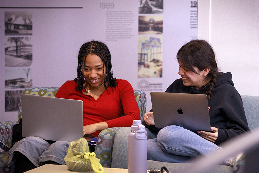 Two students interacting sit next to each other with laptops on their laps.