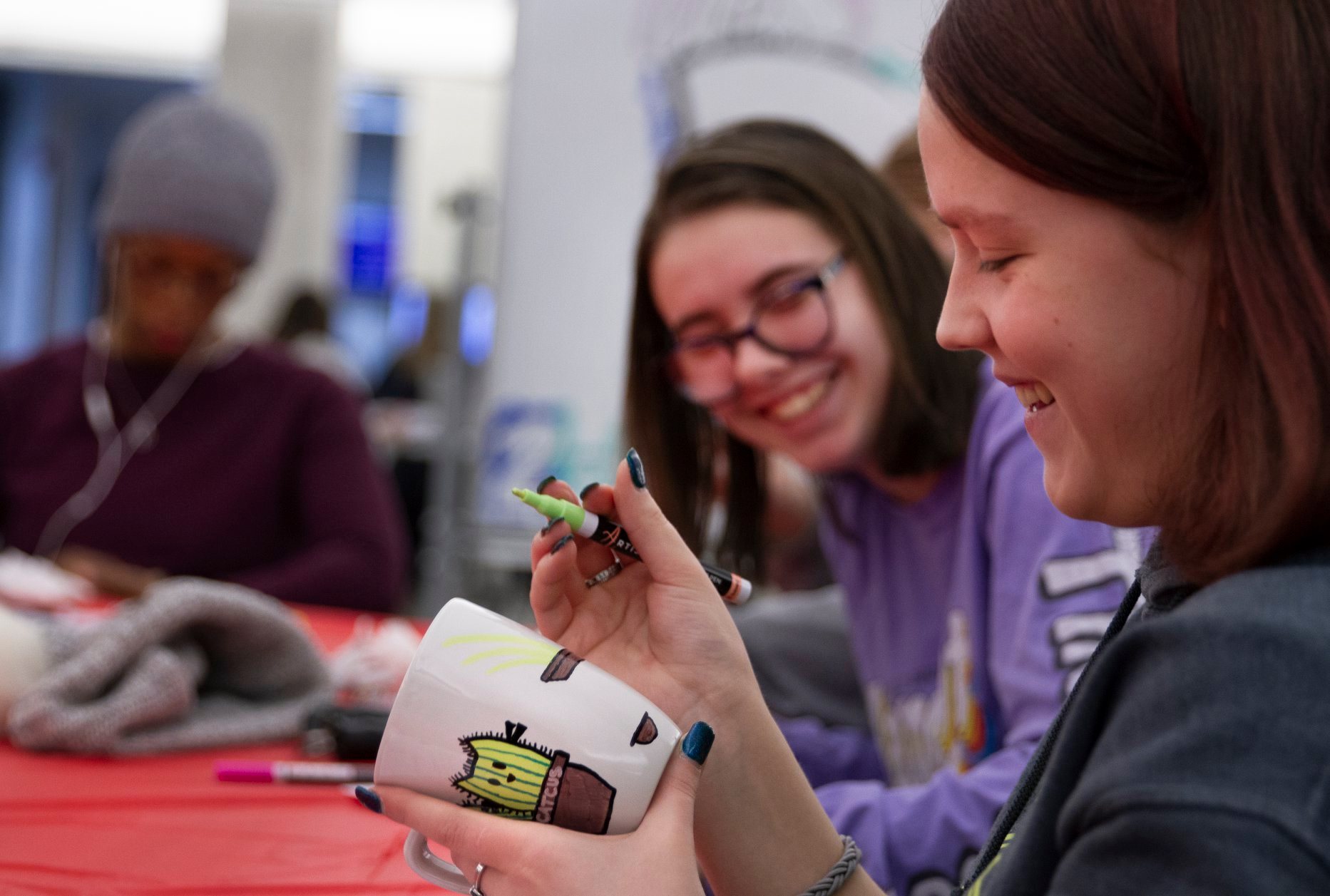 Two women sitting at a table and decorating mugs while laughing.